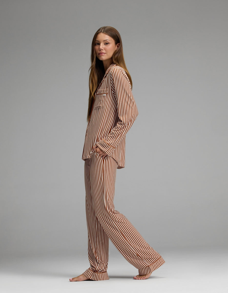 A person stands barefoot, wearing the Cozy Earth Women’s Bamboo Stretch-Knit Long Sleeve Pajama Set in a brown and white stripe pattern, against a plain grey background.