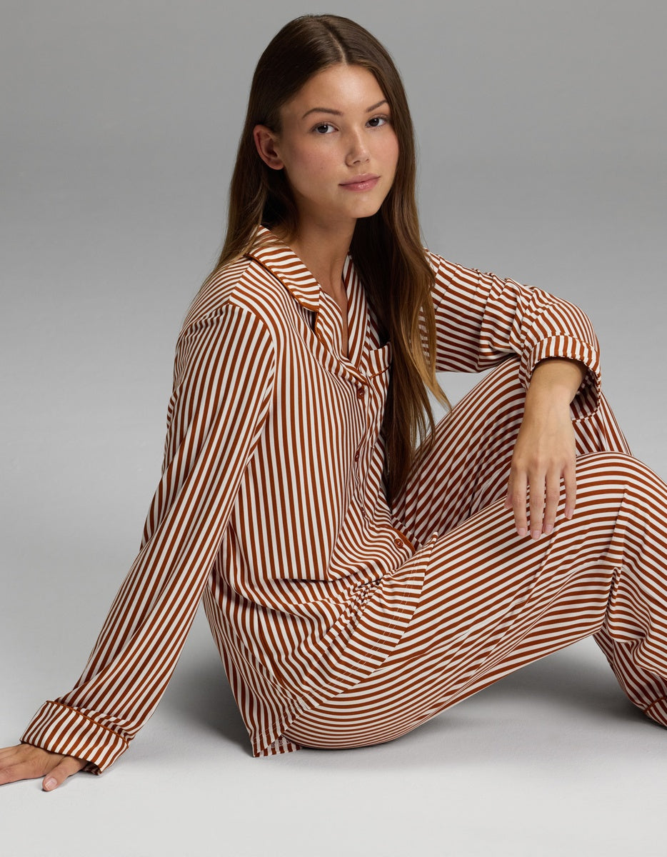 A woman with long brown hair sits on the floor against a plain gray background, wearing the Cozy Earth Women’s Bamboo Stretch-Knit Long Sleeve Pajama Set in a brown and white striped pattern, looking at the camera with a neutral expression.