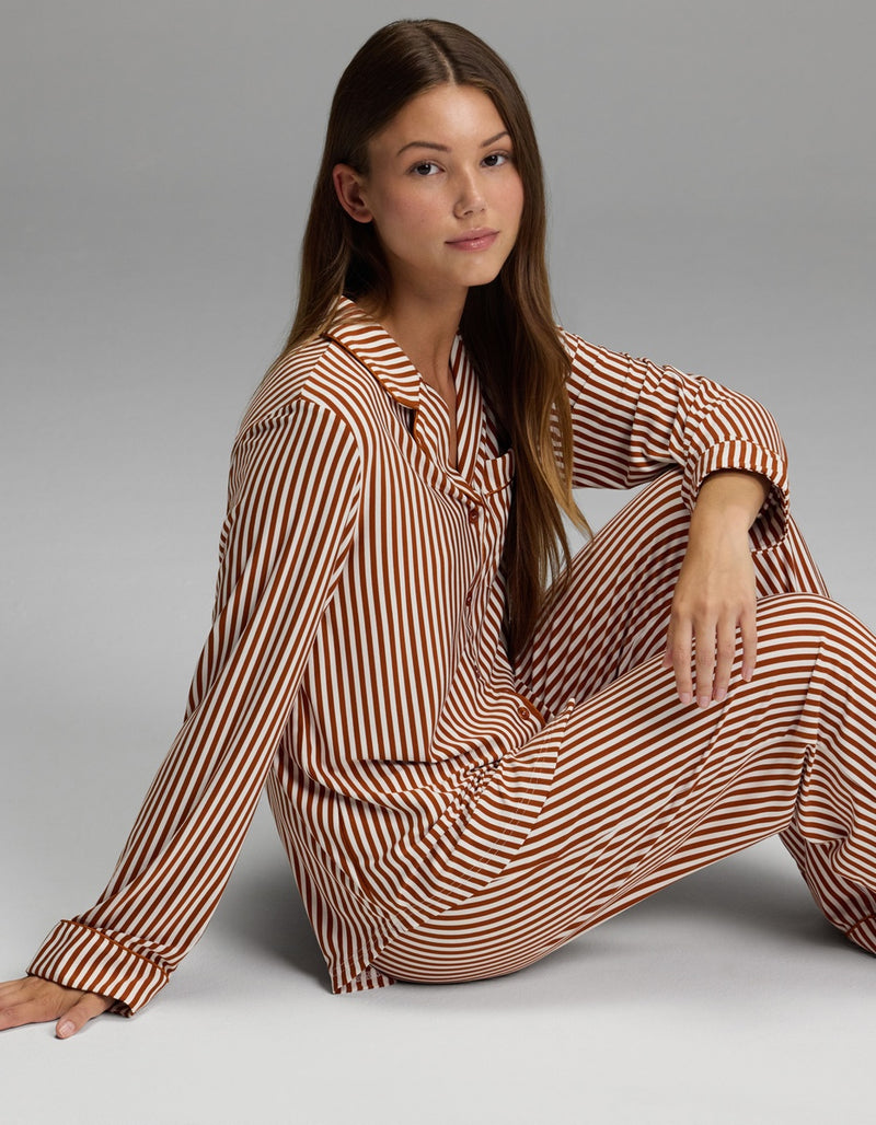 A woman with long brown hair sits on the floor against a plain gray background, wearing the Cozy Earth Women’s Bamboo Stretch-Knit Long Sleeve Pajama Set in a brown and white striped pattern, looking at the camera with a neutral expression.