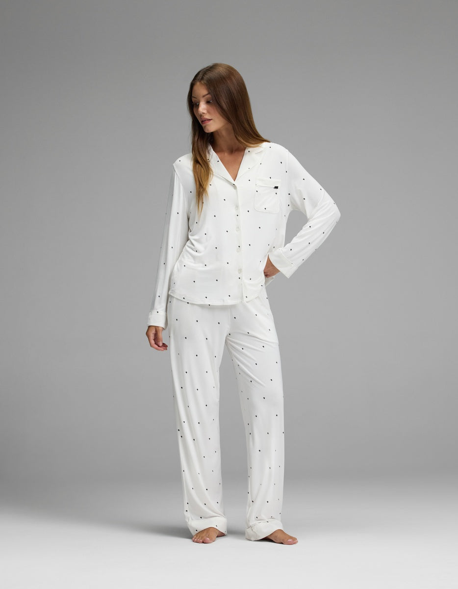 A woman models the Cozy Earth Women’s Bamboo Stretch-Knit Long Sleeve Pajama Set in white with small black polka dots, standing barefoot on a gray background and looking to her left with one hand on her hip.