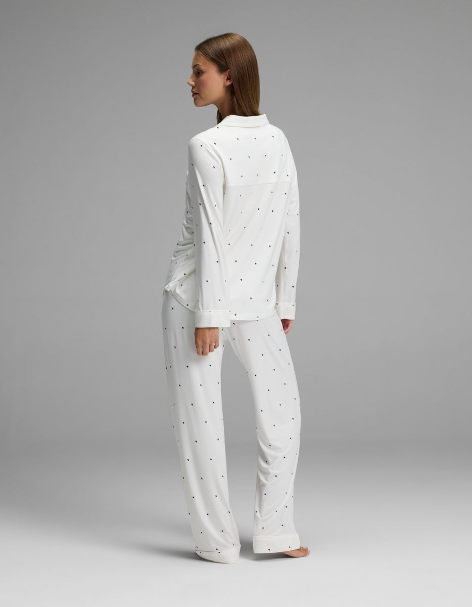 A woman stands barefoot in a plain studio setting, facing away, wearing the Cozy Earth Women’s Bamboo Stretch-Knit Long Sleeve Pajama Set in white with small dot patterns.