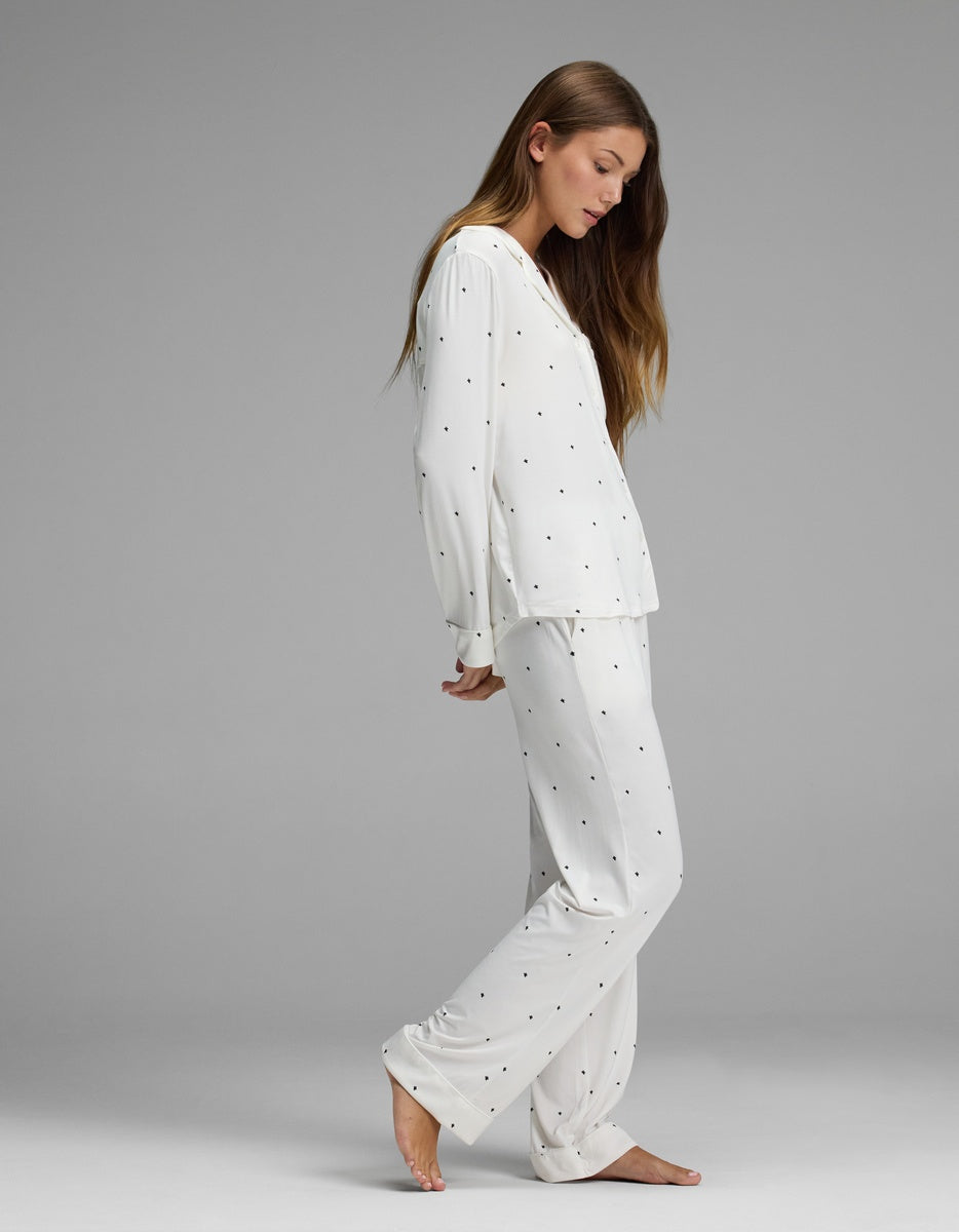 A woman models the Cozy Earth Women’s Bamboo Stretch-Knit Long Sleeve Pajama Set in white with small black dots, standing barefoot on a gray background in a side profile with her head gently tilted down and her long hair draped over one shoulder.