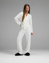 A woman wears the Cozy Earth Women’s Bamboo Stretch-Knit Long Sleeve Pajama Set in white polka dot print, paired with black slippers, as she stands in a relaxed pose against a plain gray background.|Color:Ivory Mini Ghost