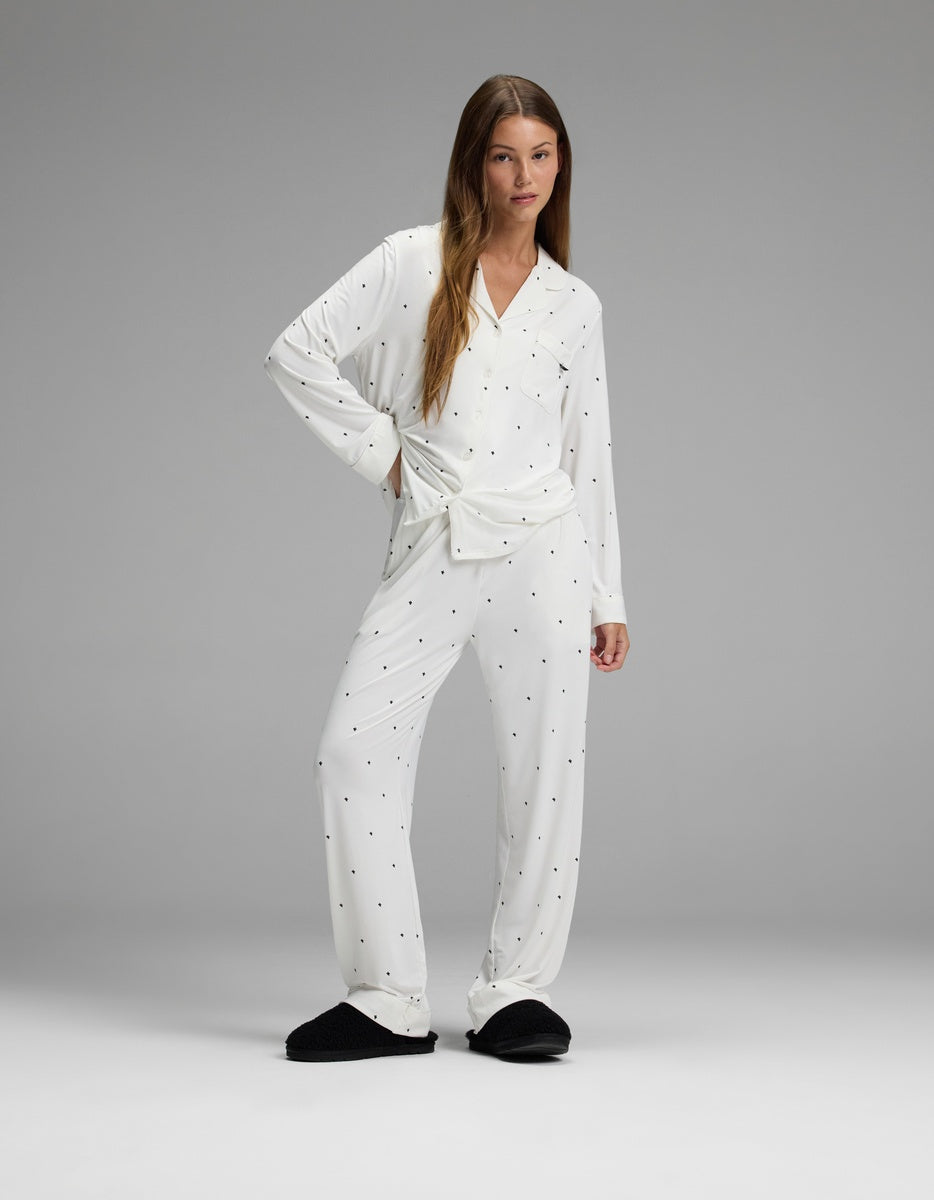 A woman wears the Cozy Earth Women’s Bamboo Stretch-Knit Long Sleeve Pajama Set in white polka dot print, paired with black slippers, as she stands in a relaxed pose against a plain gray background.