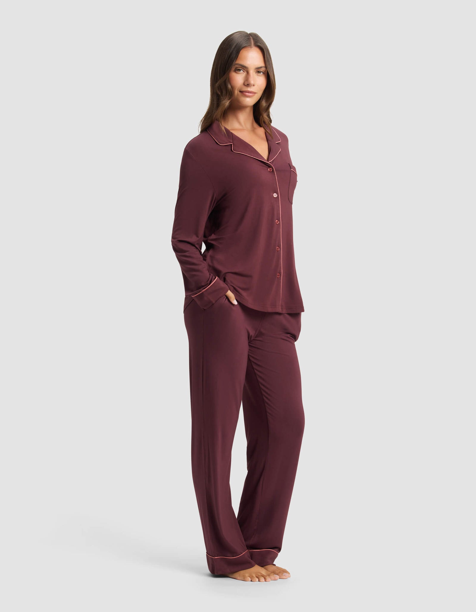 A woman stands barefoot against a light gray background, wearing the Cozy Earth Women's Bamboo Stretch-Knit Long Sleeve Pajama Set in burgundy with buttons and matching pants.