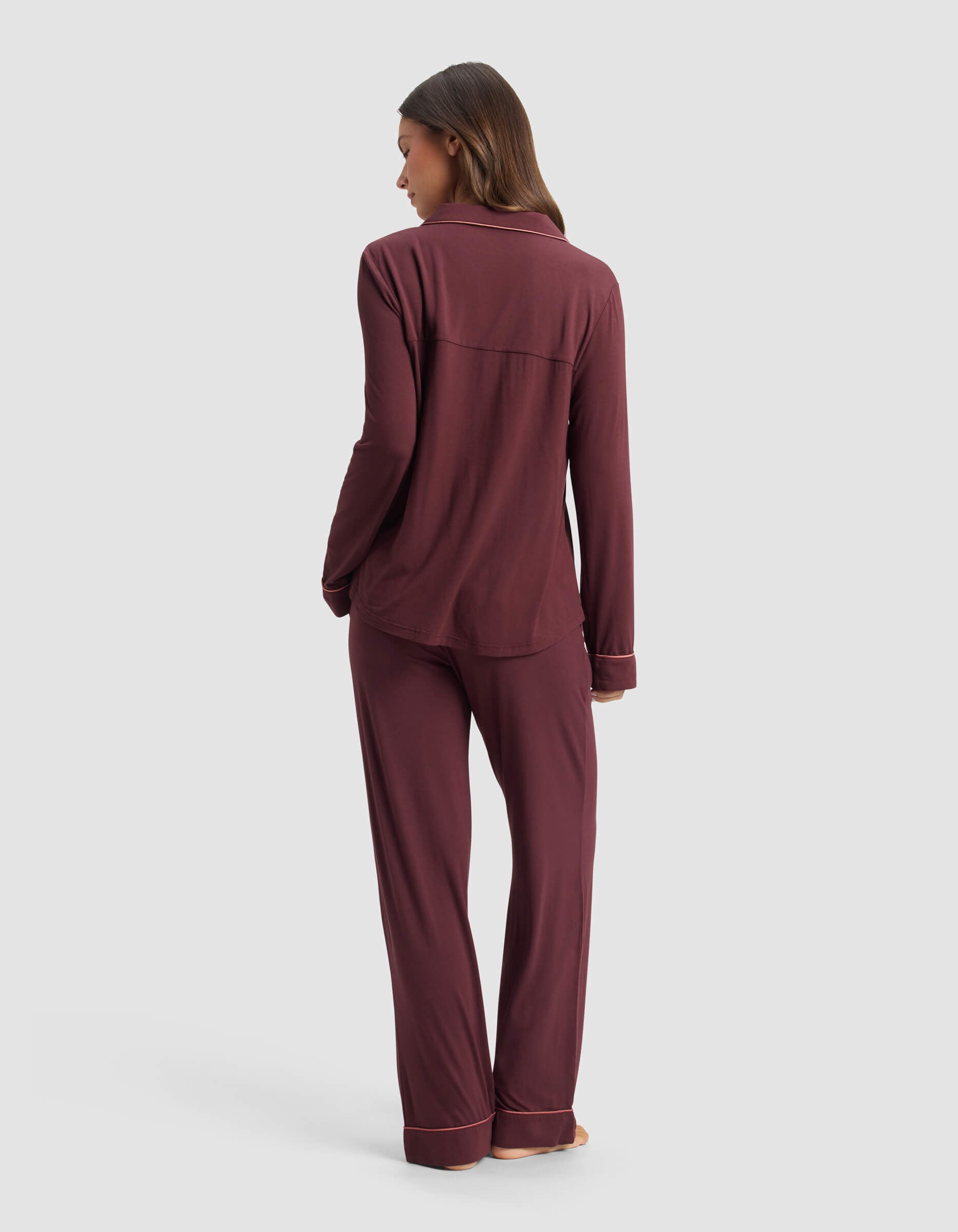 A woman stands barefoot facing away, modeling the Cozy Earth Women's Bamboo Stretch-Knit Long Sleeve Pajama Set in burgundy with matching pants against a plain light gray background. 