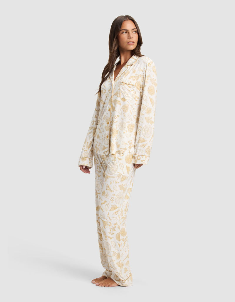 A woman stands barefoot in Cozy Earth's Women's Bamboo Stretch-Knit Long Sleeve Pajama Set, featuring a white base with a gold floral print, posing against a plain light gray background.