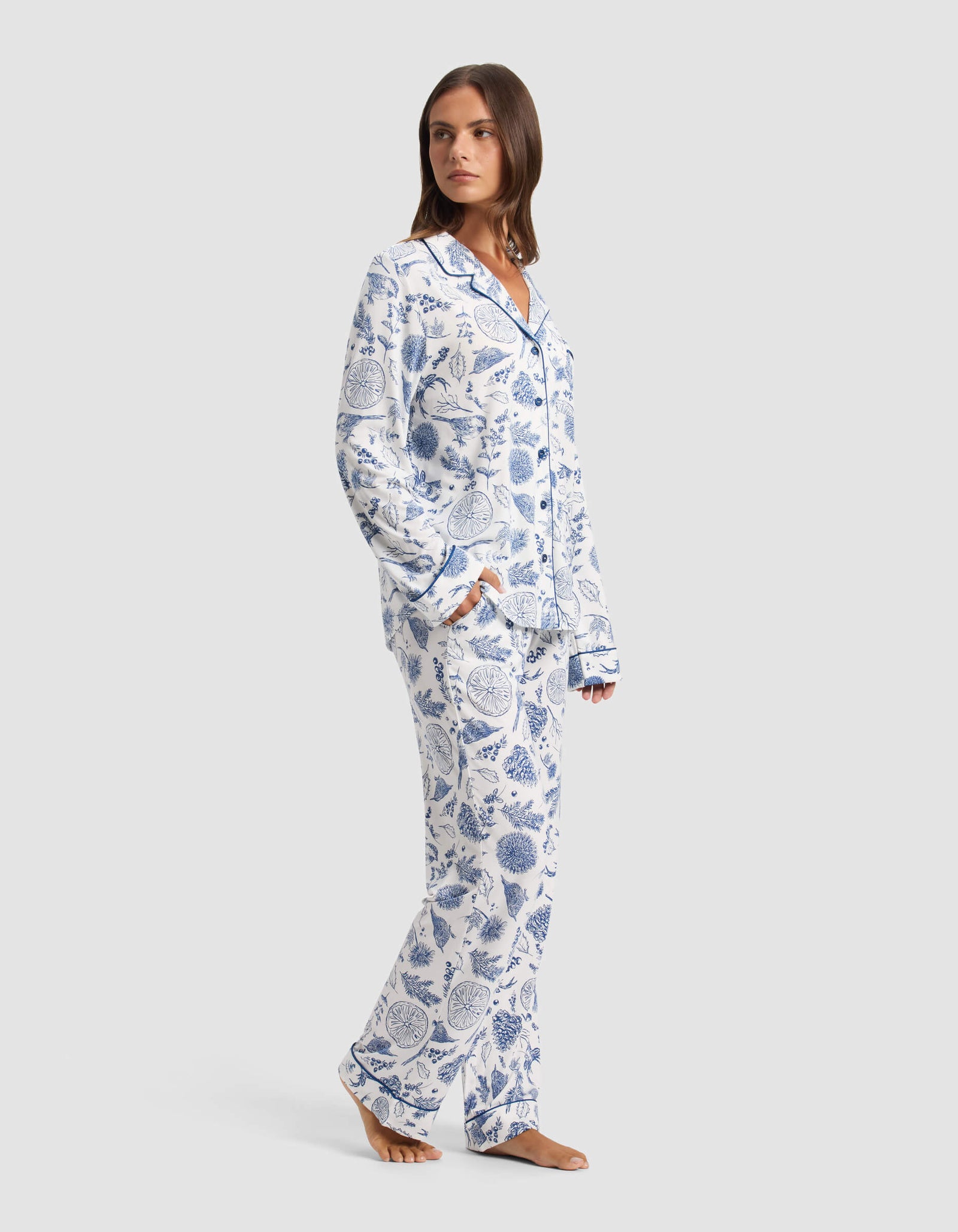 A woman stands barefoot wearing Cozy Earth's Women's Bamboo Stretch-Knit Pajama Pant, featuring a white background with blue botanical print and paired with a matching long-sleeve button-up top. She looks over her shoulder. 