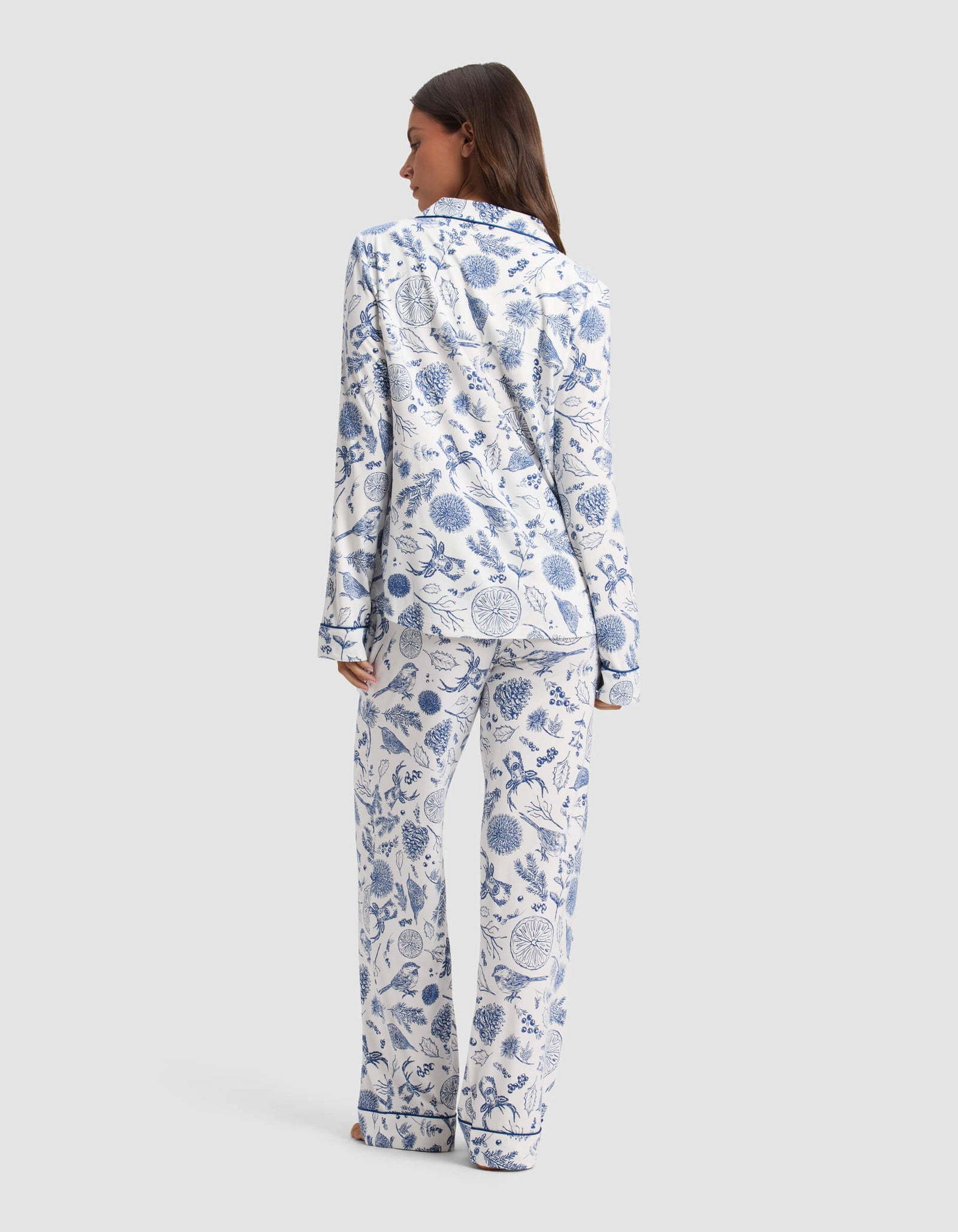 A woman stands barefoot facing away, wearing the Cozy Earth Women's Bamboo Stretch-Knit Long Sleeve Pajama Set in a blue and white botanical print against a plain light gray background.