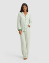 A woman stands barefoot in Cozy Earth’s Women's Bamboo Stretch-Knit Long Sleeve Pajama Set - Market Stripe in Divi Green, featuring light green and white vertical stripes, buttoned top with chest pocket, and matching pants. She looks neutral with a hand in her pocket.
