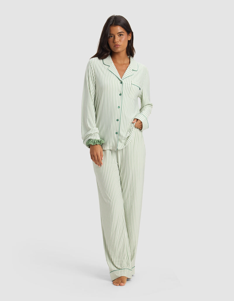 A woman stands barefoot in Cozy Earth’s Women's Bamboo Stretch-Knit Long Sleeve Pajama Set - Market Stripe in Divi Green, featuring light green and white vertical stripes, buttoned top with chest pocket, and matching pants. She looks neutral with a hand in her pocket.