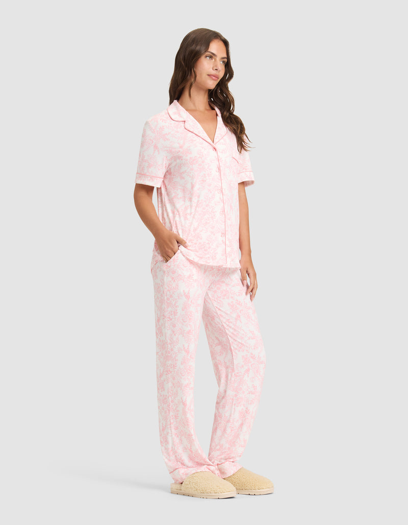 A woman wears the Cozy Earth Women's Bamboo Stretch-Knit Long Sleeve Pajama Set and beige slippers, standing with hands in her pockets and looking to the side.
