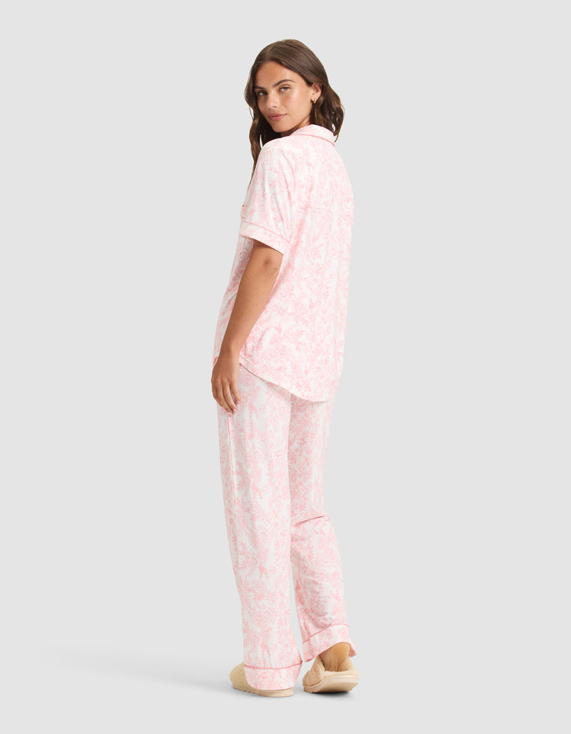 A woman, facing away and glancing over her shoulder, wears the Cozy Earth Women's Bamboo Stretch-Knit Long Sleeve Pajama Set in light pink and white with a floral pattern and beige slippers against a plain light gray background.