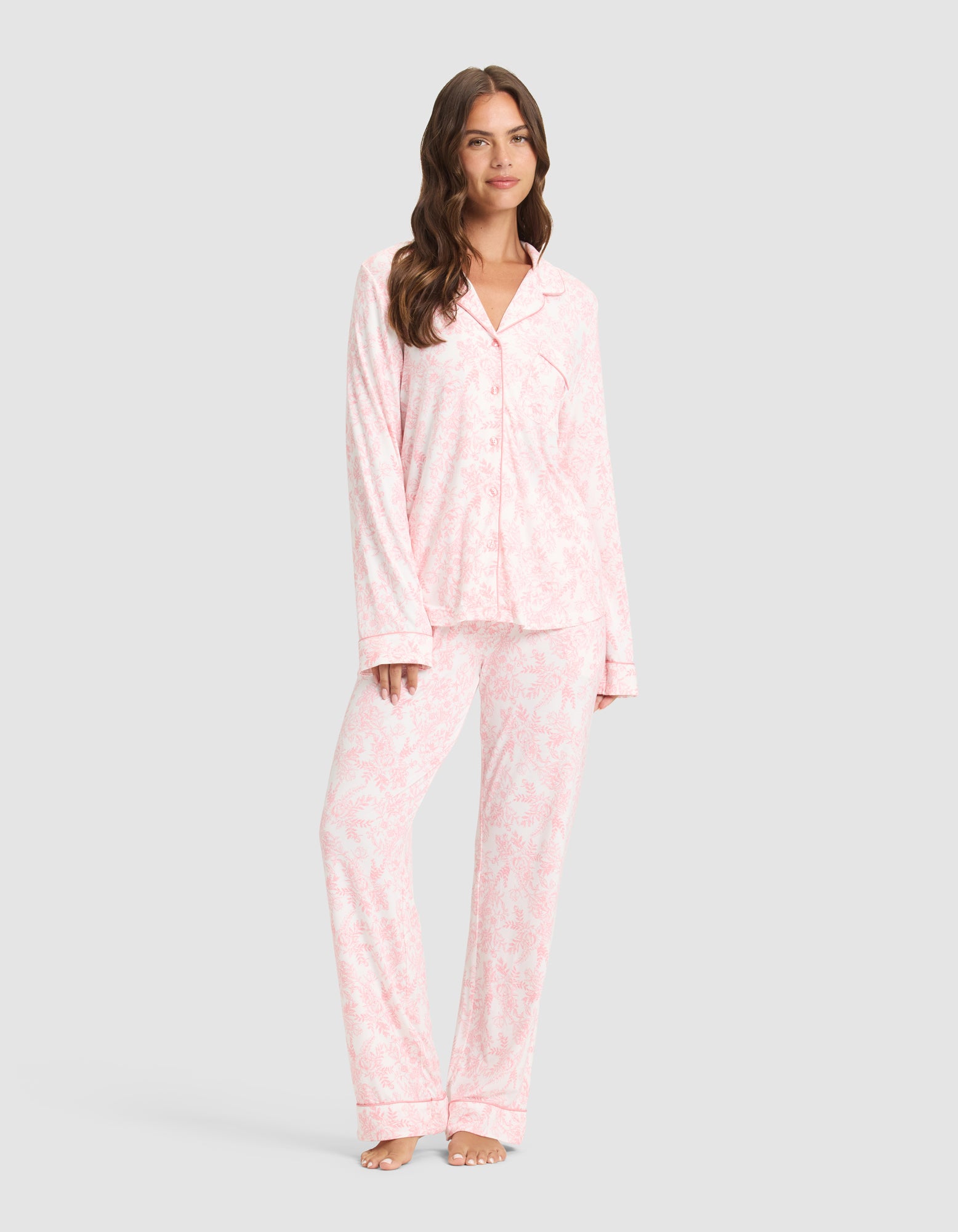 A woman with long, wavy brown hair smiles slightly while wearing the Cozy Earth Women's Bamboo Stretch-Knit Long Sleeve Pajama Set, featuring a matching light pink and white button-up top and loose pants, standing barefoot against a plain background. |Color:Tulip Toile