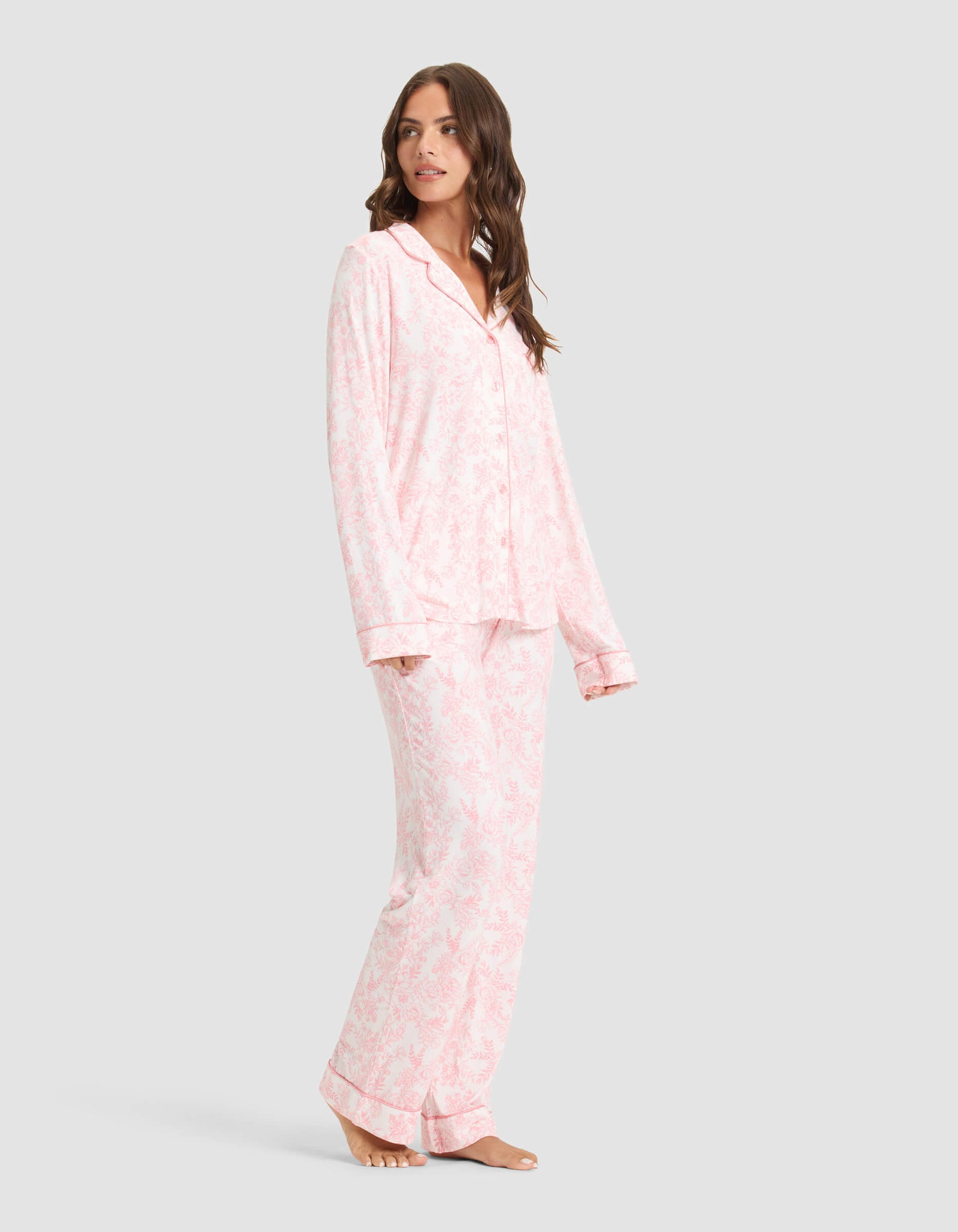 A woman poses barefoot against a light gray background wearing Cozy Earth's Women's Bamboo Stretch-Knit Long Sleeve Pajama Set in a light pink and white pattern, featuring a button-up top and matching pants. She looks over her shoulder. 