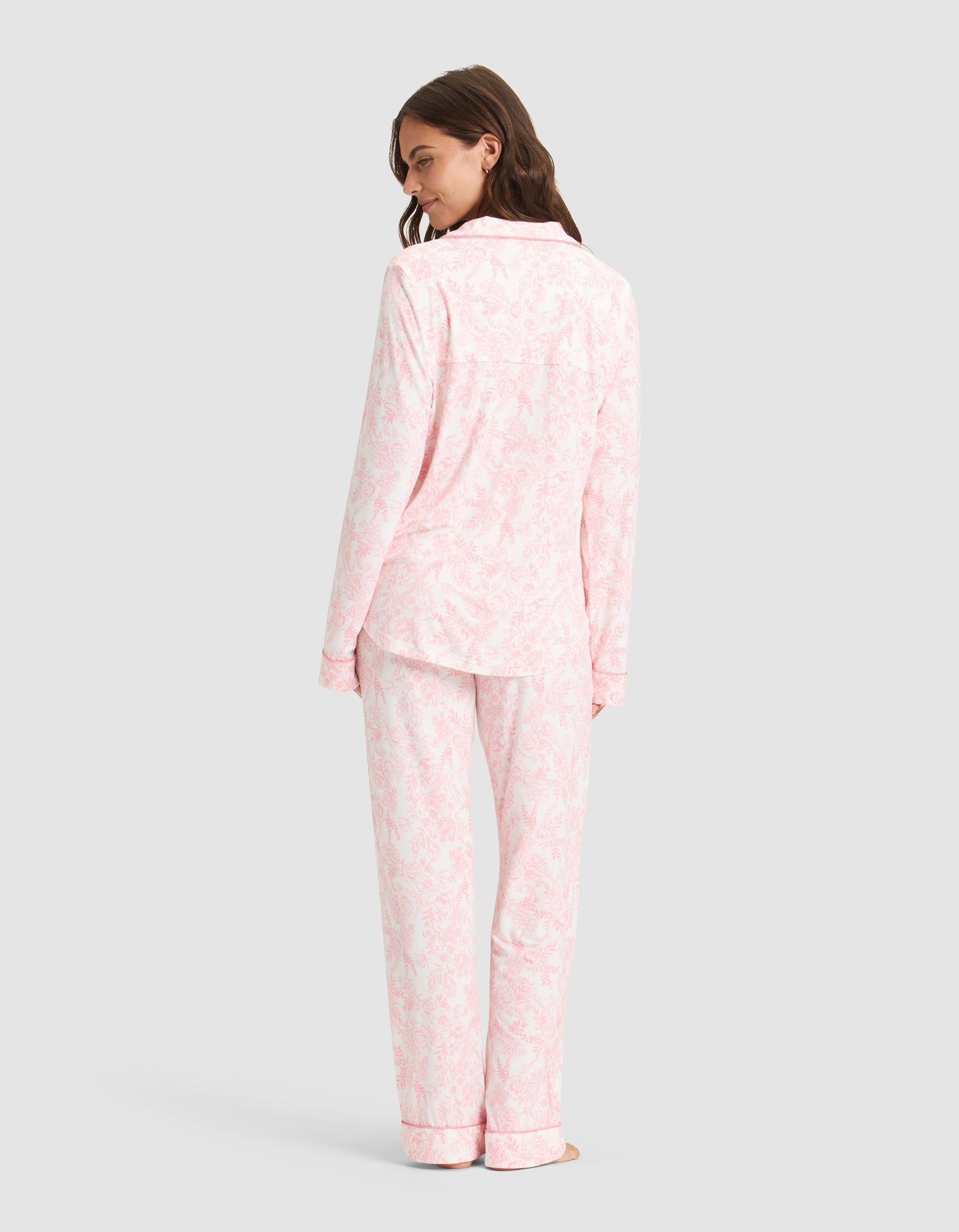A woman stands barefoot, facing away, wearing the Cozy Earth Women's Bamboo Stretch-Knit Long Sleeve Pajama Set in light pink and white pattern. She looks over her shoulder and smiles gently against a plain light gray background. 