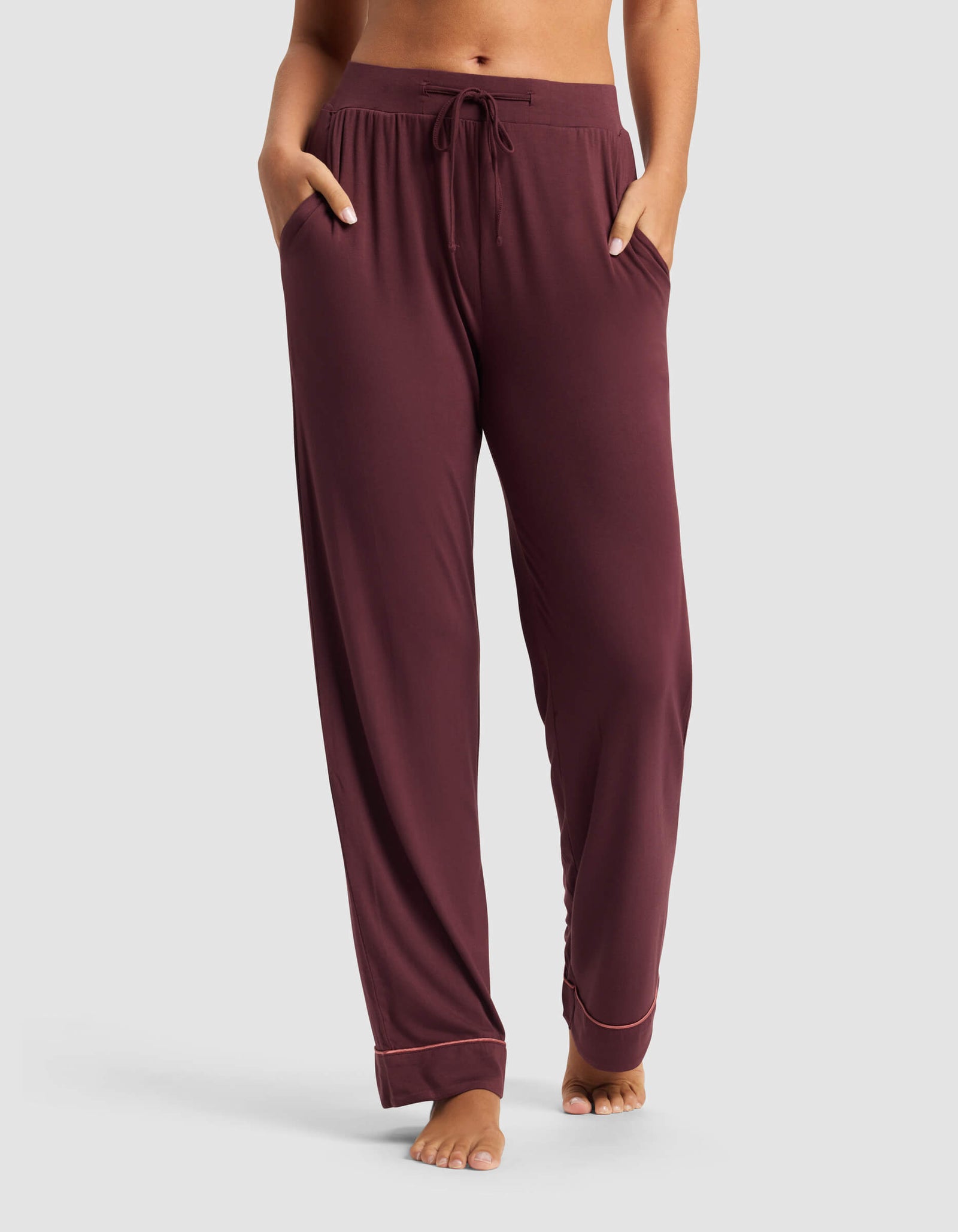 A woman wears Cozy Earth's Women's Bamboo Stretch-Knit Pajama Pant in burgundy, featuring a drawstring waist and pockets, standing barefoot against a plain gray background.
