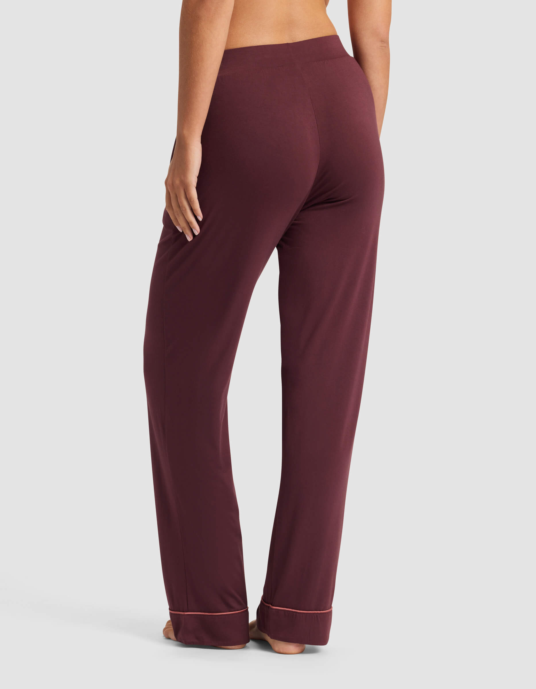 A person stands barefoot on a white background, wearing Cozy Earth's Women's Bamboo Stretch-Knit Pajama Pant in burgundy with a relaxed fit and light-trimmed cuffs; their back and legs are visible from mid-back to feet.|Color:Burgundy