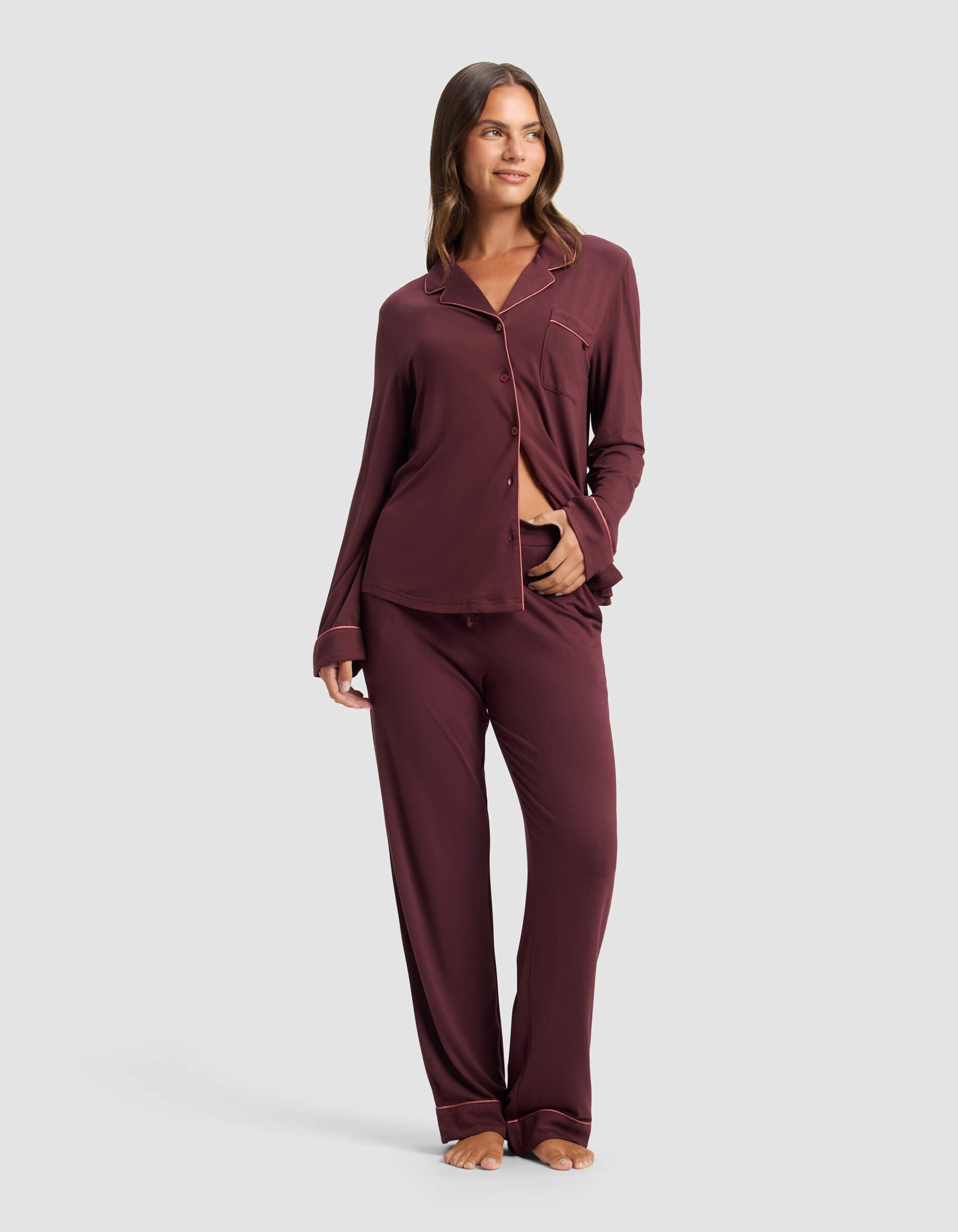 A woman stands barefoot, smiling with one hand on her waist, wearing the Cozy Earth Women's Bamboo Stretch-Knit Long Sleeve Pajama Set—a matching maroon outfit featuring a collared, button-up top with a chest pocket and matching pants. 
