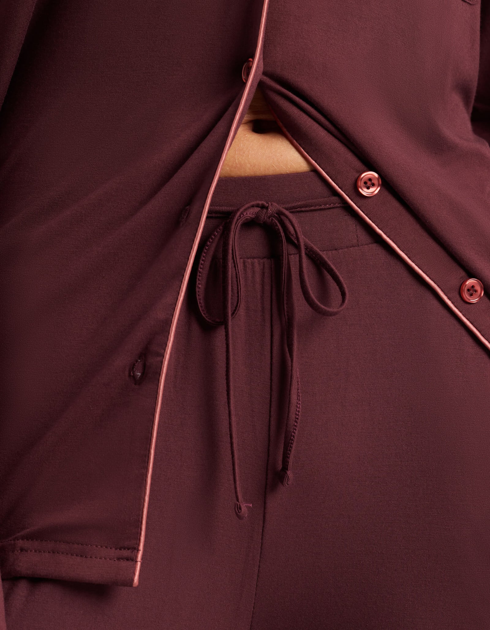 Close-up of a person in Cozy Earth's Women's Bamboo Stretch-Knit Pajama Pant, featuring burgundy fabric with pink piping accents and an elastic drawstring waistband shown beneath a partially unbuttoned pajama top.