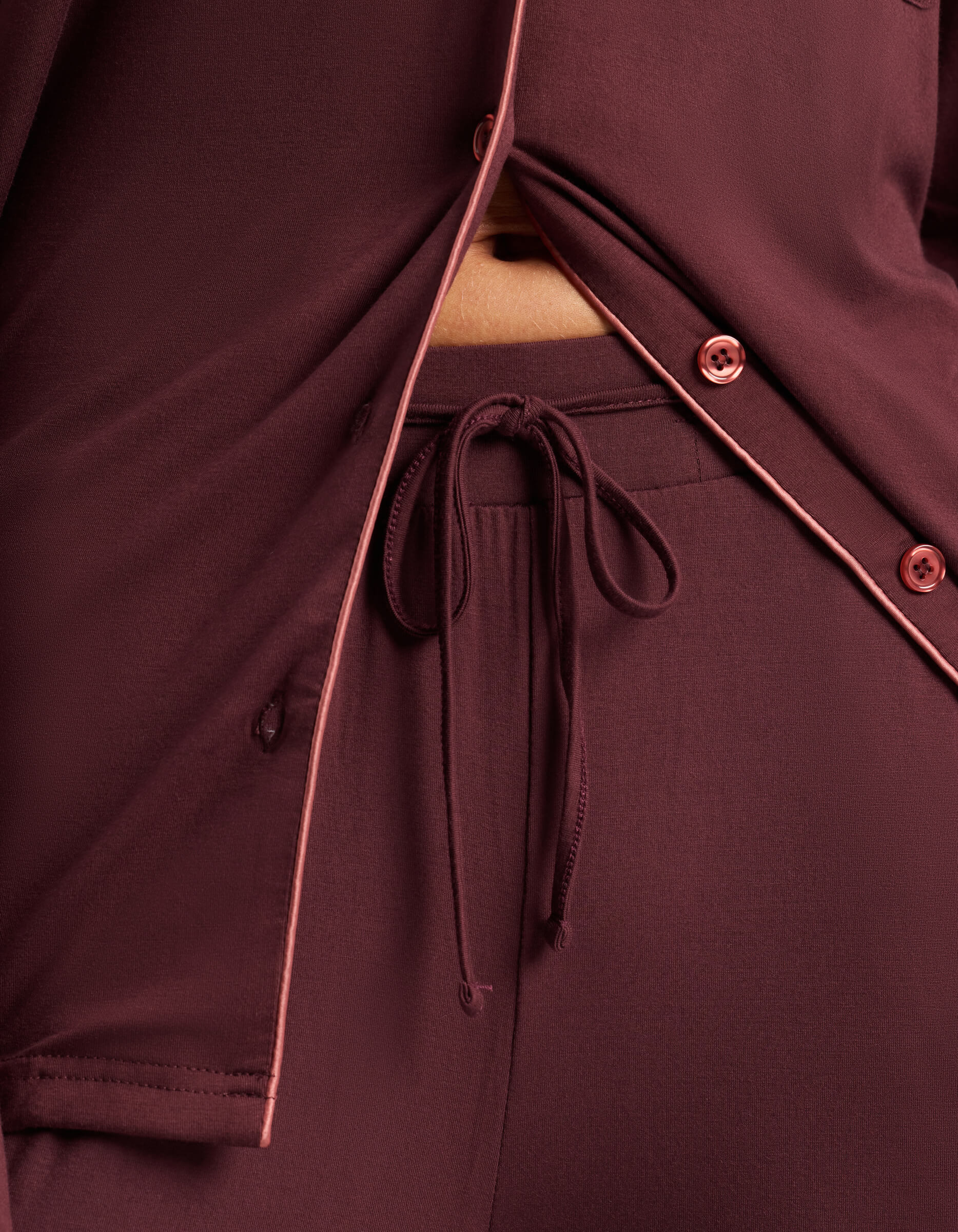 Close-up of a person in Cozy Earth's Women's Bamboo Stretch-Knit Pajama Pant, featuring burgundy fabric with pink piping accents and an elastic drawstring waistband shown beneath a partially unbuttoned pajama top.