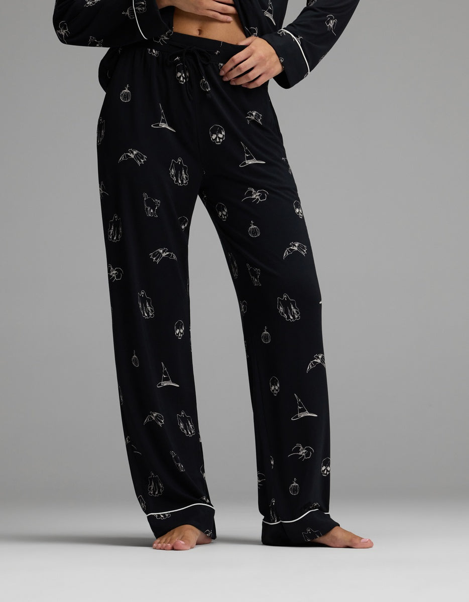 A person stands barefoot against a gray background, wearing the Cozy Earth Women’s Bamboo Stretch-Knit Long Sleeve Pajama Set in black, featuring white Halloween-themed prints of bats, pumpkins, skulls, and witch hats.