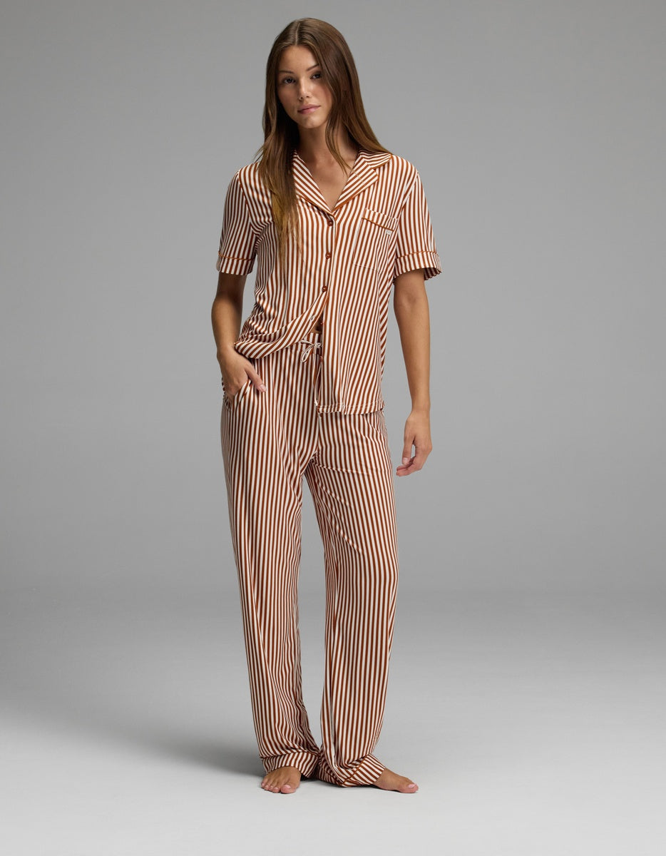 A woman stands barefoot against a plain gray background, wearing Cozy Earth’s Women’s Bamboo Stretch-Knit Pajama Pant in a matching set with brown and white vertical stripes, featuring short sleeves and long pants. Her right hand is in her pocket as she looks slightly toward the camera.