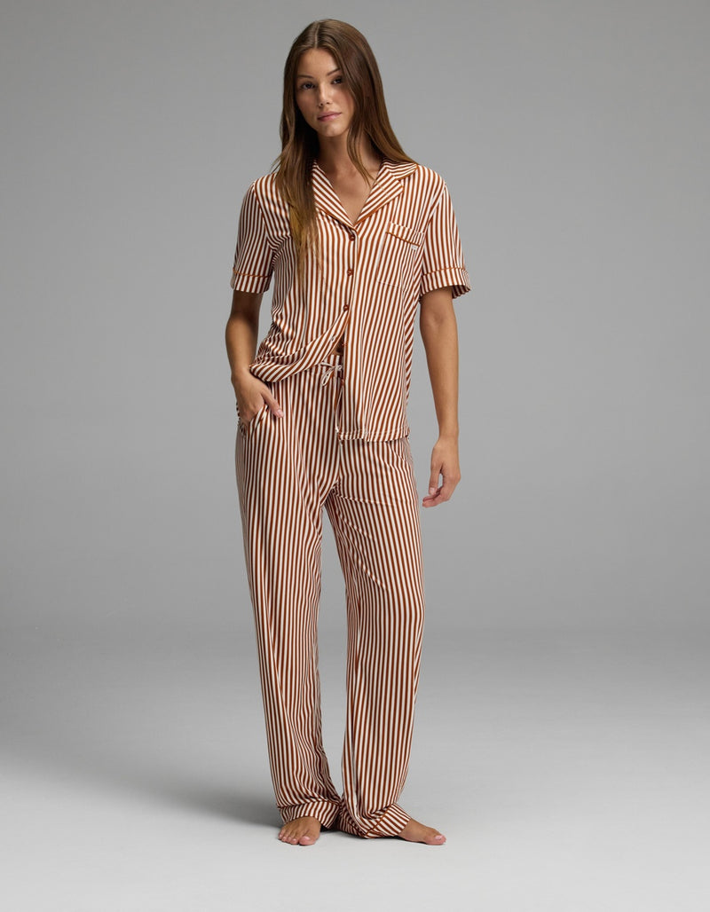 A woman stands barefoot against a plain gray background, wearing Cozy Earth’s Women’s Bamboo Stretch-Knit Pajama Pant in a matching set with brown and white vertical stripes, featuring short sleeves and long pants. Her right hand is in her pocket as she looks slightly toward the camera.