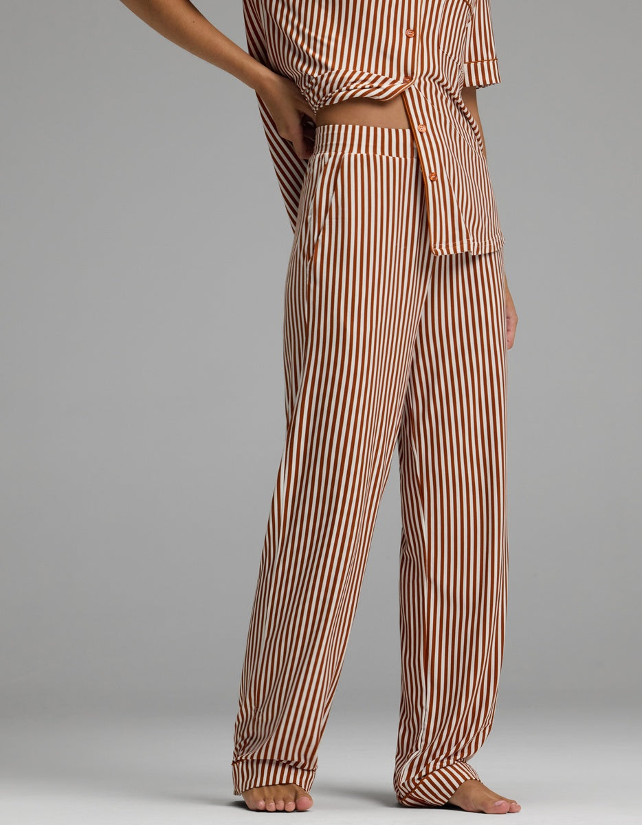 A person stands barefoot on a plain gray background, wearing the Cozy Earth Women’s Bamboo Stretch-Knit Pajama Pant in a brown and white vertical striped pattern with the matching top, cropped at the chest.|Color:Market Stripe in Cinnamon