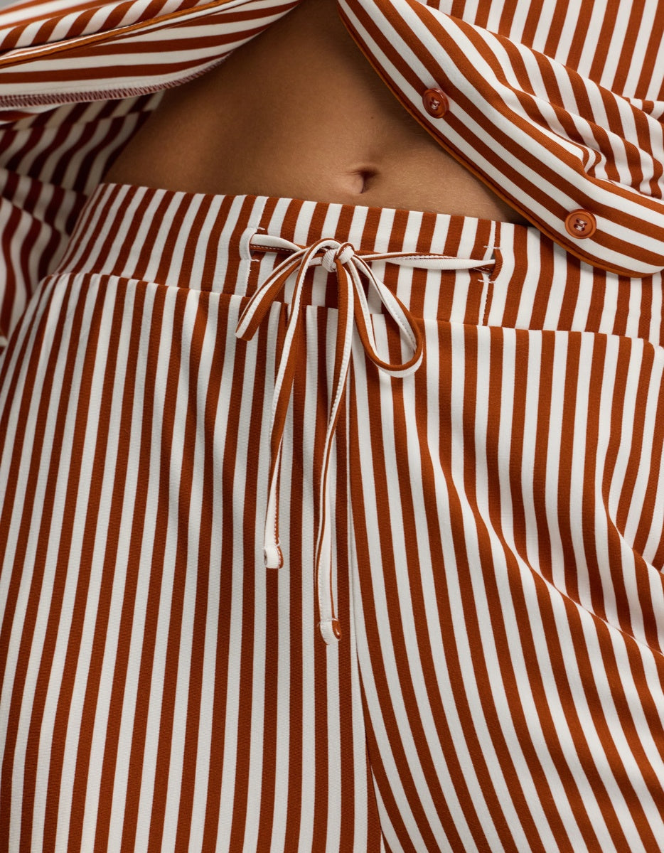 Close-up of a person’s midsection wearing the Cozy Earth Women’s Bamboo Stretch-Knit Pajama Pant, featuring brown and white vertical stripes with a drawstring waist, paired with a partially open matching shirt that reveals their belly button.