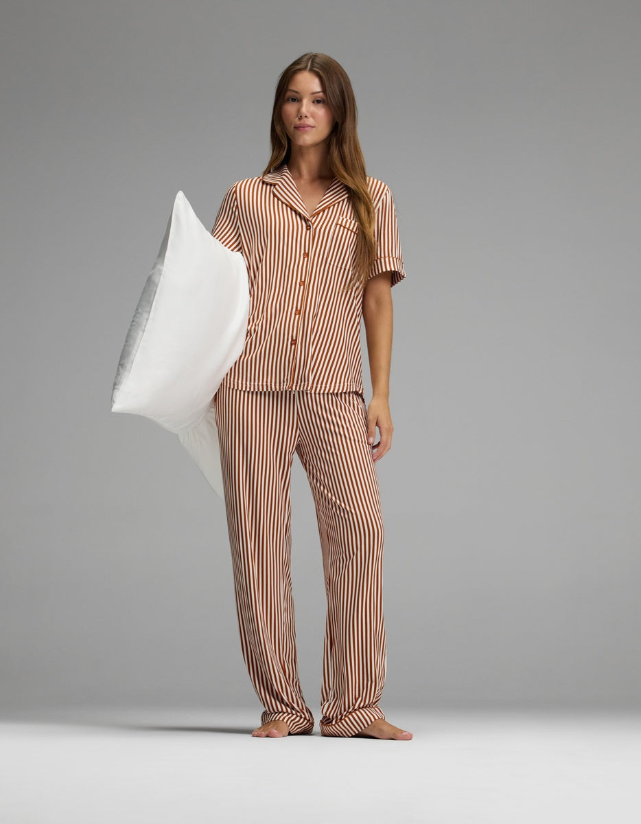 A woman stands barefoot on a gray floor, wearing Cozy Earth’s Women’s Bamboo Stretch-Knit Pajama Pant in an orange-and-white striped pattern and holding a large white pillow under her left arm. The background is plain and gray with evenly soft lighting.
