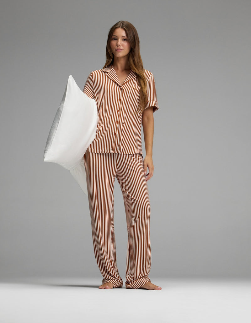 A woman stands barefoot on a gray floor, wearing Cozy Earth’s Women’s Bamboo Stretch-Knit Pajama Pant in an orange-and-white striped pattern and holding a large white pillow under her left arm. The background is plain and gray with evenly soft lighting.