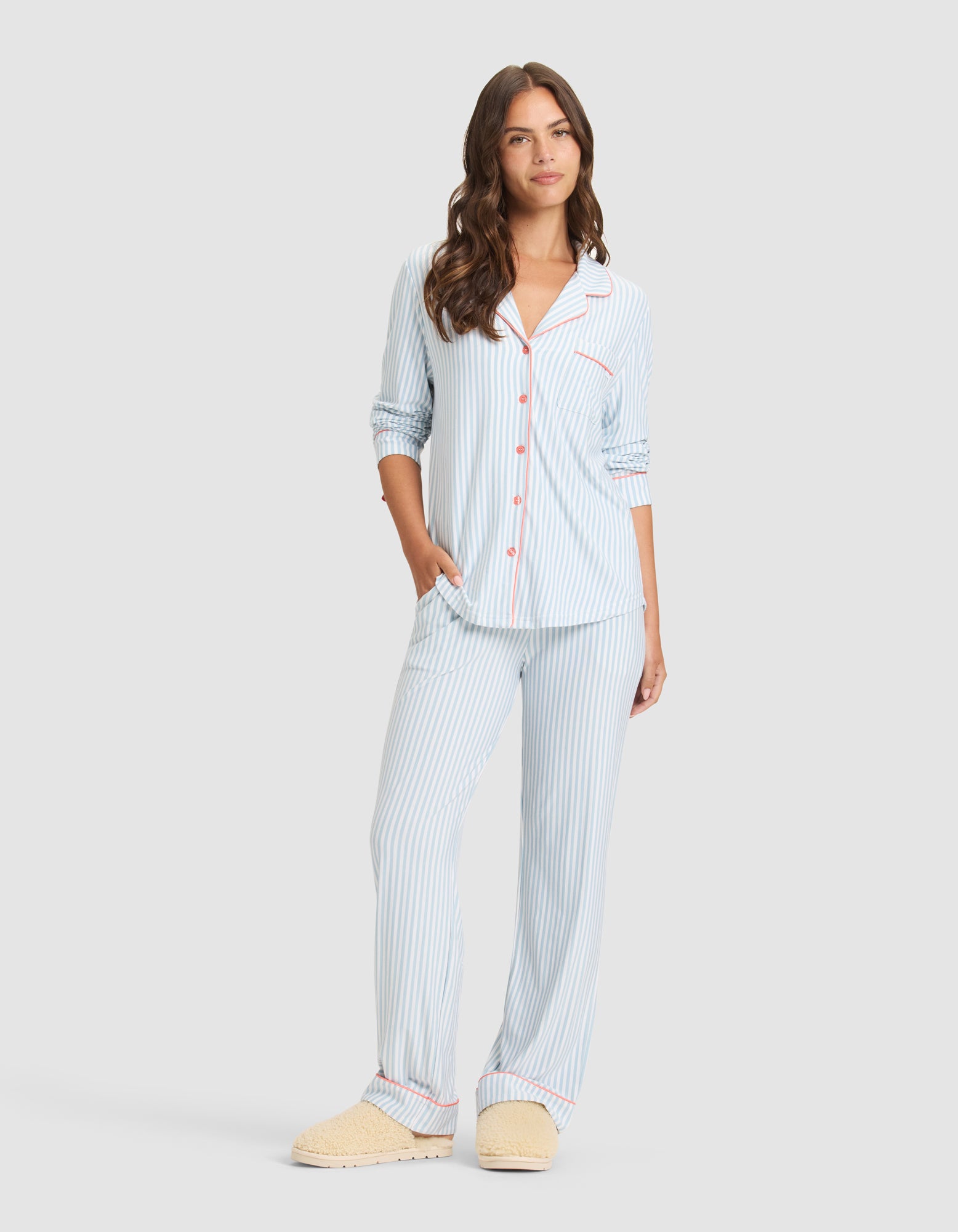 A woman wears the Cozy Earth Women's Bamboo Stretch-Knit Long Sleeve Pajama Set in light blue and white stripes with red piping and beige slippers. The top features buttons, a chest pocket, and she stands with one hand in her pocket. 