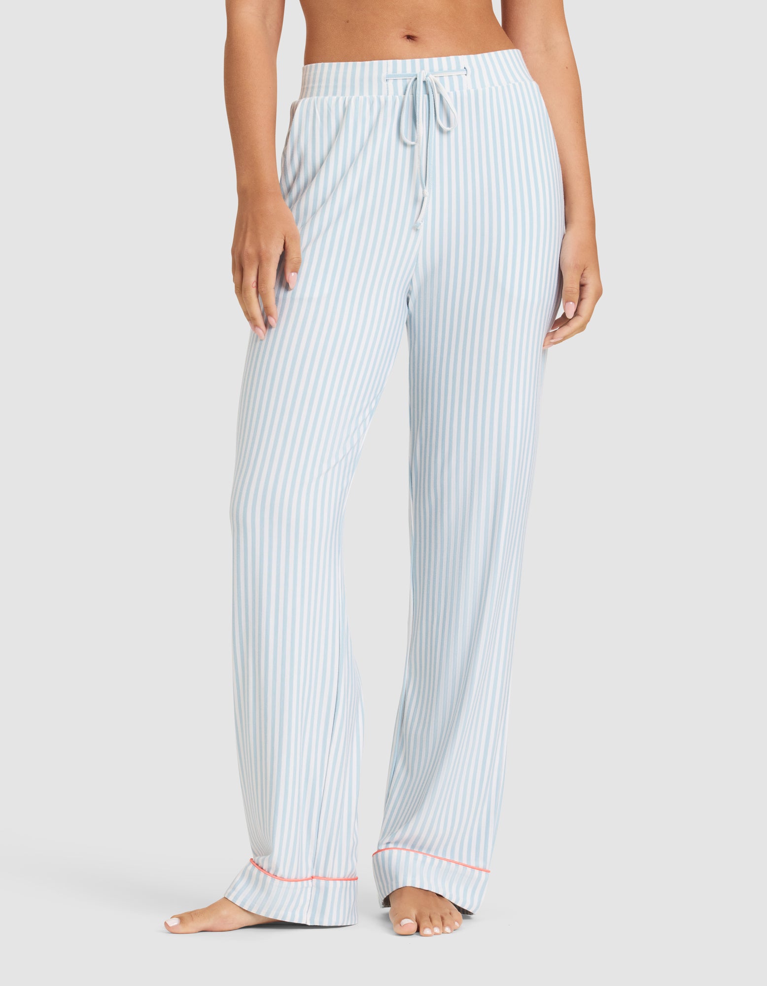 A woman models the Cozy Earth Women's Bamboo Stretch-Knit Long Sleeve Pajama Set in light blue and white vertical stripes with a drawstring waist, loose fit, and red trim at the cuffs, standing barefoot against a plain background. 