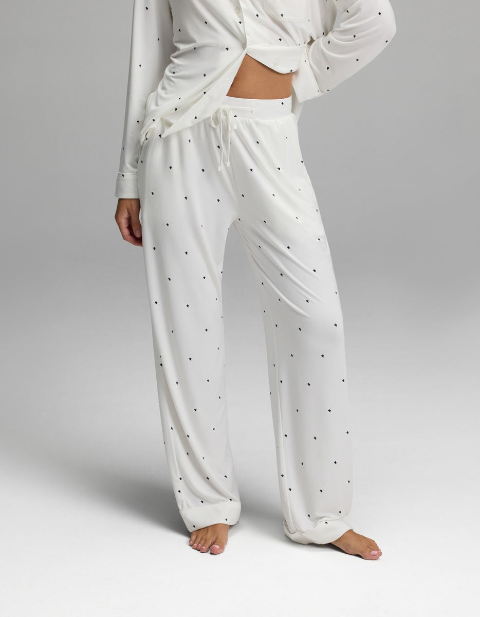 A person is wearing the Cozy Earth Women’s Bamboo Stretch-Knit Long Sleeve Pajama Set, which features a white fabric with small black polka dots, a long-sleeve top, and drawstring pants. The image displays the outfit from shoulders to feet against a plain background.