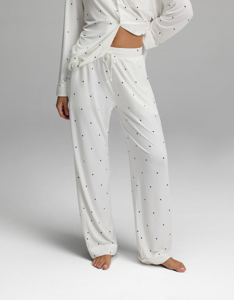 A person is wearing the Cozy Earth Women’s Bamboo Stretch-Knit Long Sleeve Pajama Set, which features a white fabric with small black polka dots, a long-sleeve top, and drawstring pants. The image displays the outfit from shoulders to feet against a plain background.