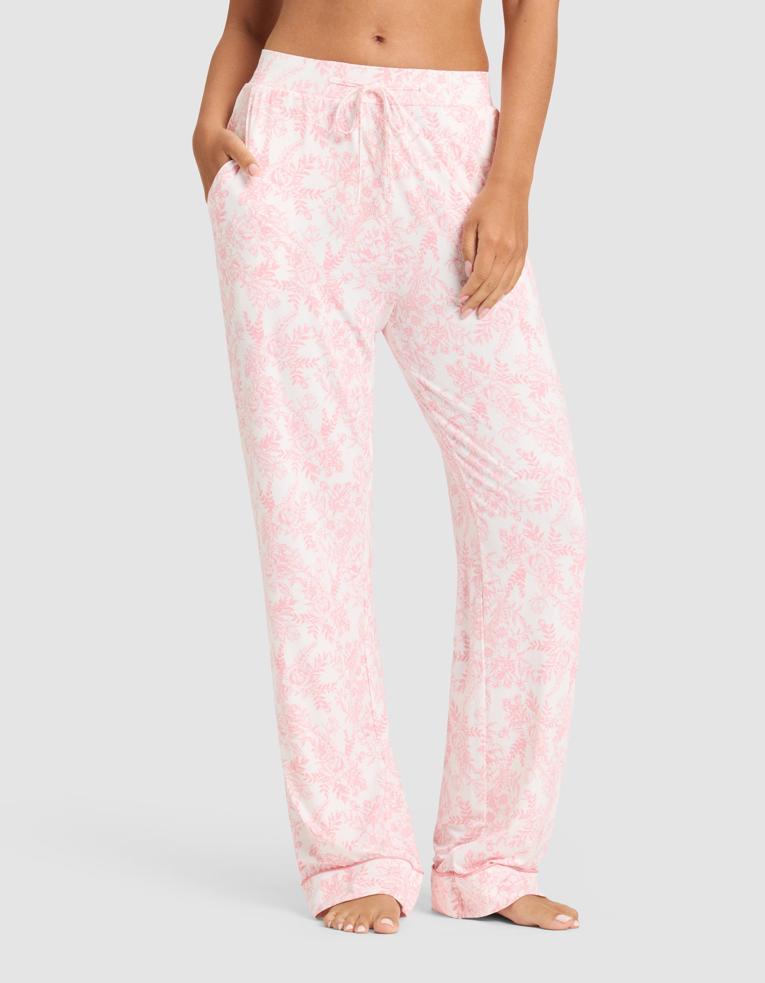 A woman wears Cozy Earth’s Women's Bamboo Stretch-Knit Long Sleeve Pajama Set in light pink and white floral, featuring relaxed-fit pants with pockets and a drawstring waist, standing barefoot against a plain background. |Color:Tulip Toile