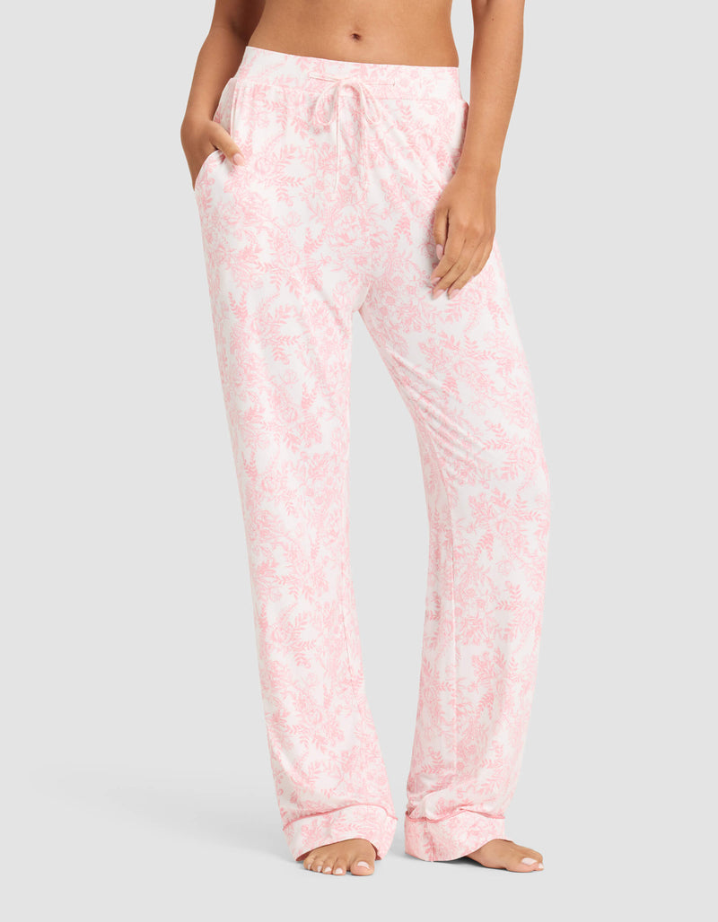 A woman wears Cozy Earth’s Women's Bamboo Stretch-Knit Long Sleeve Pajama Set in light pink and white floral, featuring relaxed-fit pants with pockets and a drawstring waist, standing barefoot against a plain background.