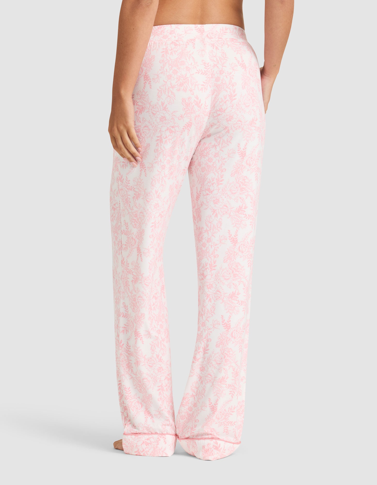 A person stands barefoot facing away, wearing Cozy Earth Women's Bamboo Stretch-Knit Long Sleeve Pajama Set in light pink and white floral. The loose-fitting pajama pants have a relaxed style against a plain light gray background. 