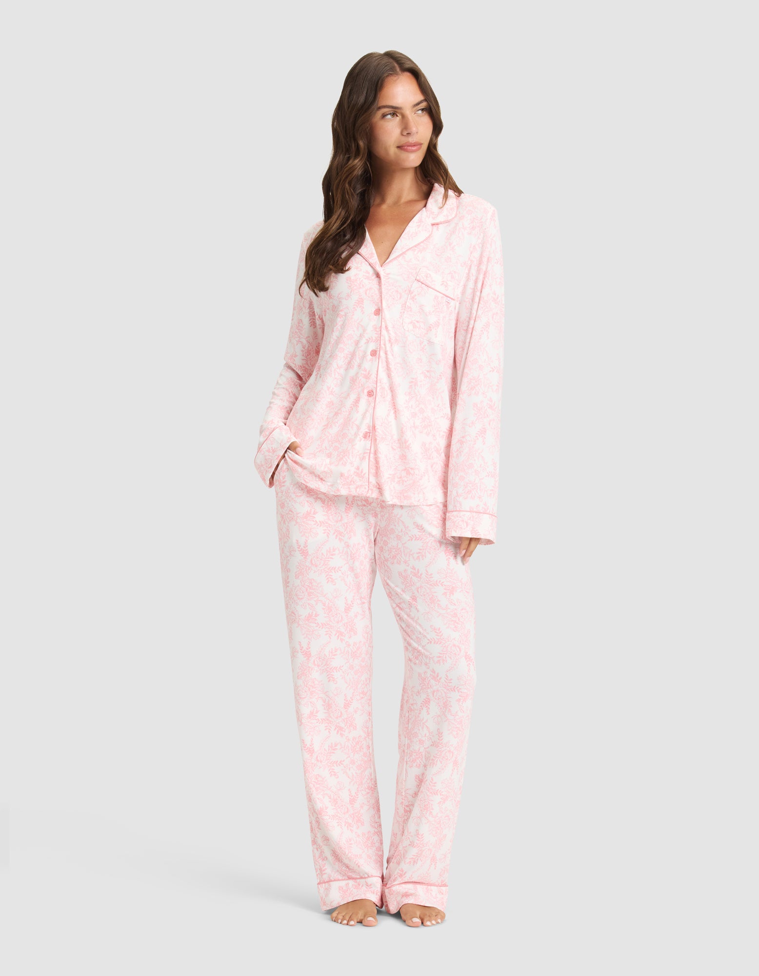 A woman poses barefoot against a light gray background wearing Cozy Earth's Women's Bamboo Stretch-Knit Long Sleeve Pajama Set in a light pink and white pattern, featuring a button-up top and matching pants. 