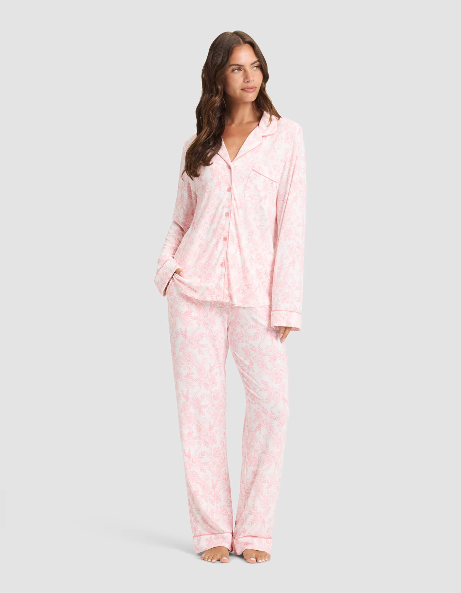 A woman poses barefoot against a light gray background wearing Cozy Earth's Women's Bamboo Stretch-Knit Long Sleeve Pajama Set in a light pink and white pattern, featuring a button-up top and matching pants. 