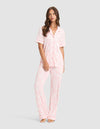A woman stands barefoot in Cozy Earth's Women's Bamboo Stretch-Knit Long Sleeve Pajama Set, featuring light pink, long pants and a matching button-up top with a subtle floral pattern, against a light gray background. |Color:Tulip Toile