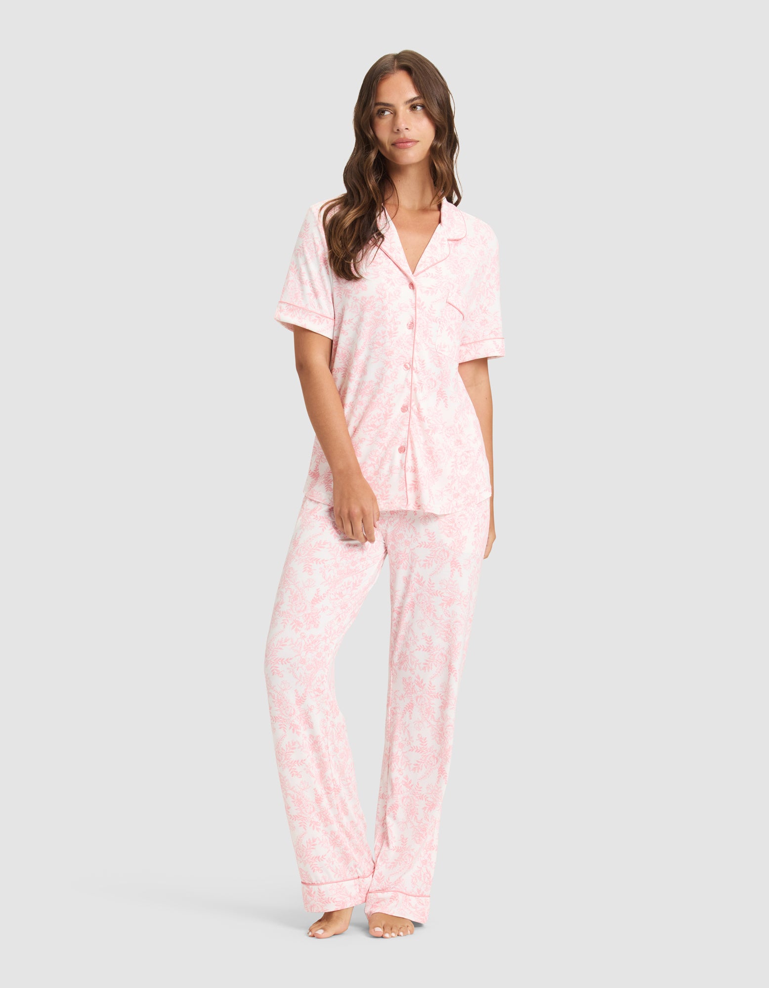 A woman stands barefoot in Cozy Earth's Women's Bamboo Stretch-Knit Long Sleeve Pajama Set, featuring light pink, long pants and a matching button-up top with a subtle floral pattern, against a light gray background. 