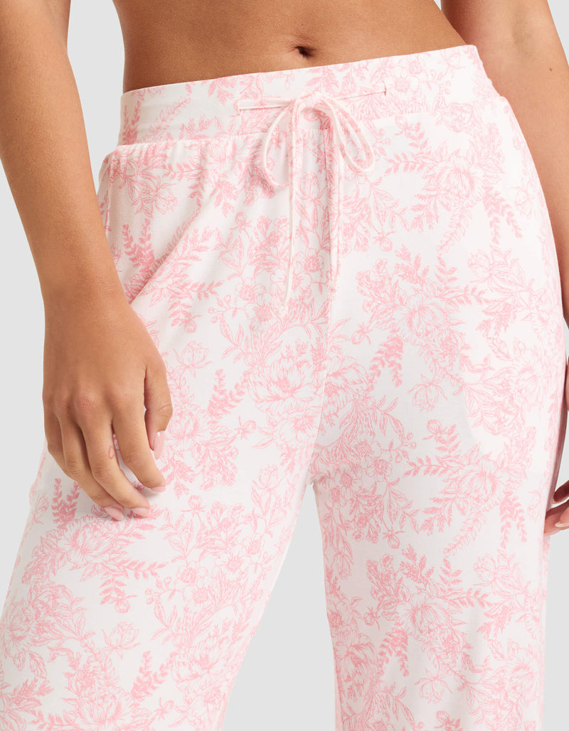A woman wears Cozy Earth's Women's Bamboo Stretch-Knit Long Sleeve Pajama Set in light pink floral, featuring drawstring pajama pants. Only her lower torso and one hand are shown against a neutral background.