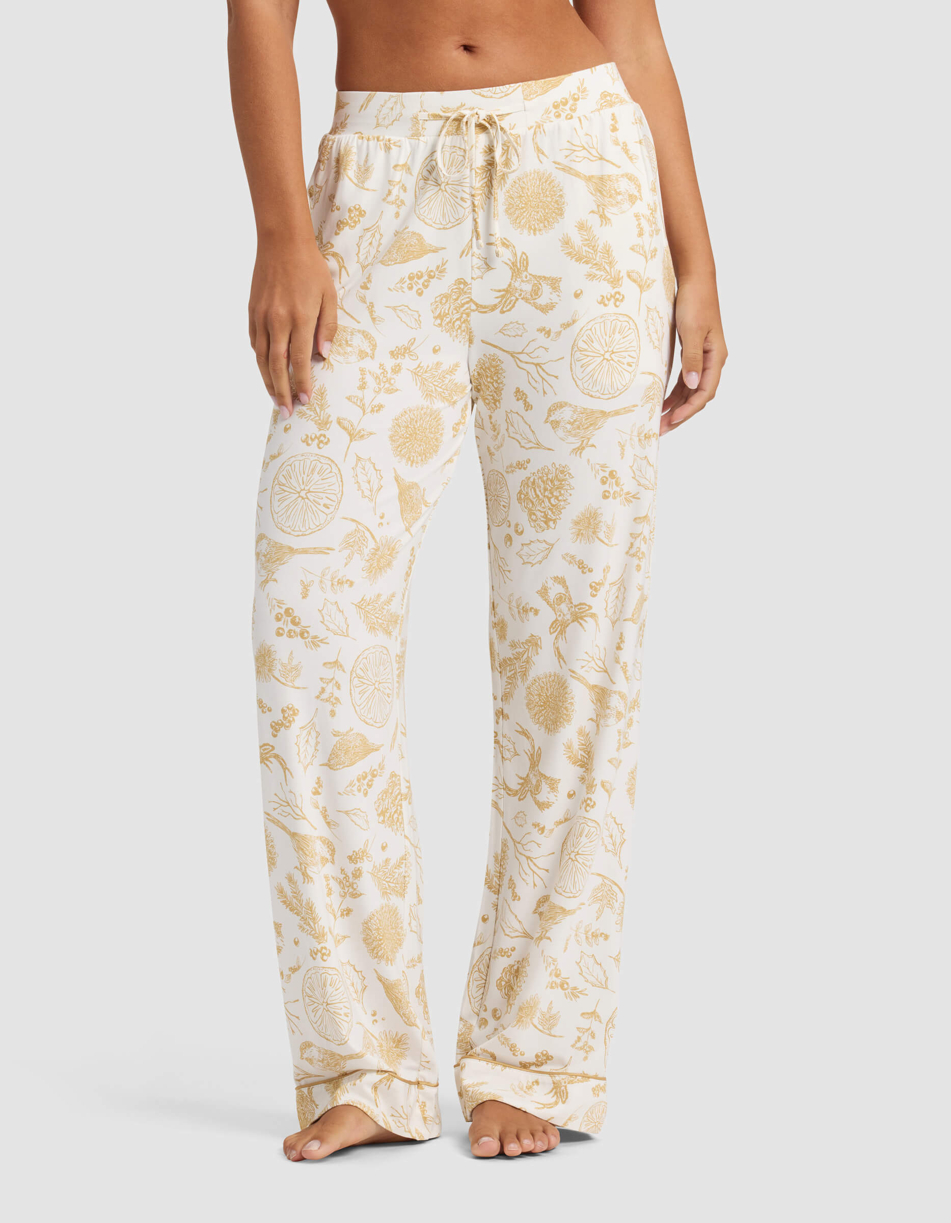 A woman wears Cozy Earth's Women's Bamboo Stretch-Knit Pajama Pant in a loose cream style with yellow floral and citrus print, standing barefoot against a plain light background.|Color:Winter Toile Dijon