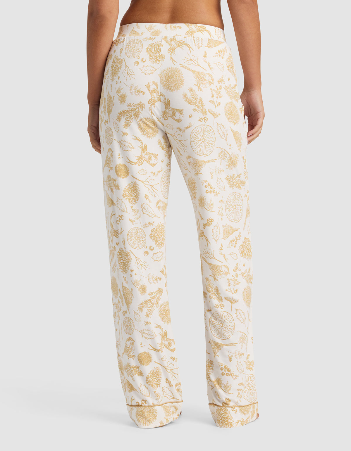 A person models the back view of the Cozy Earth Women's Bamboo Stretch-Knit Pajama Pant, featuring a gold botanical and citrus fruit print on white, shown in a relaxed fit against a plain background.