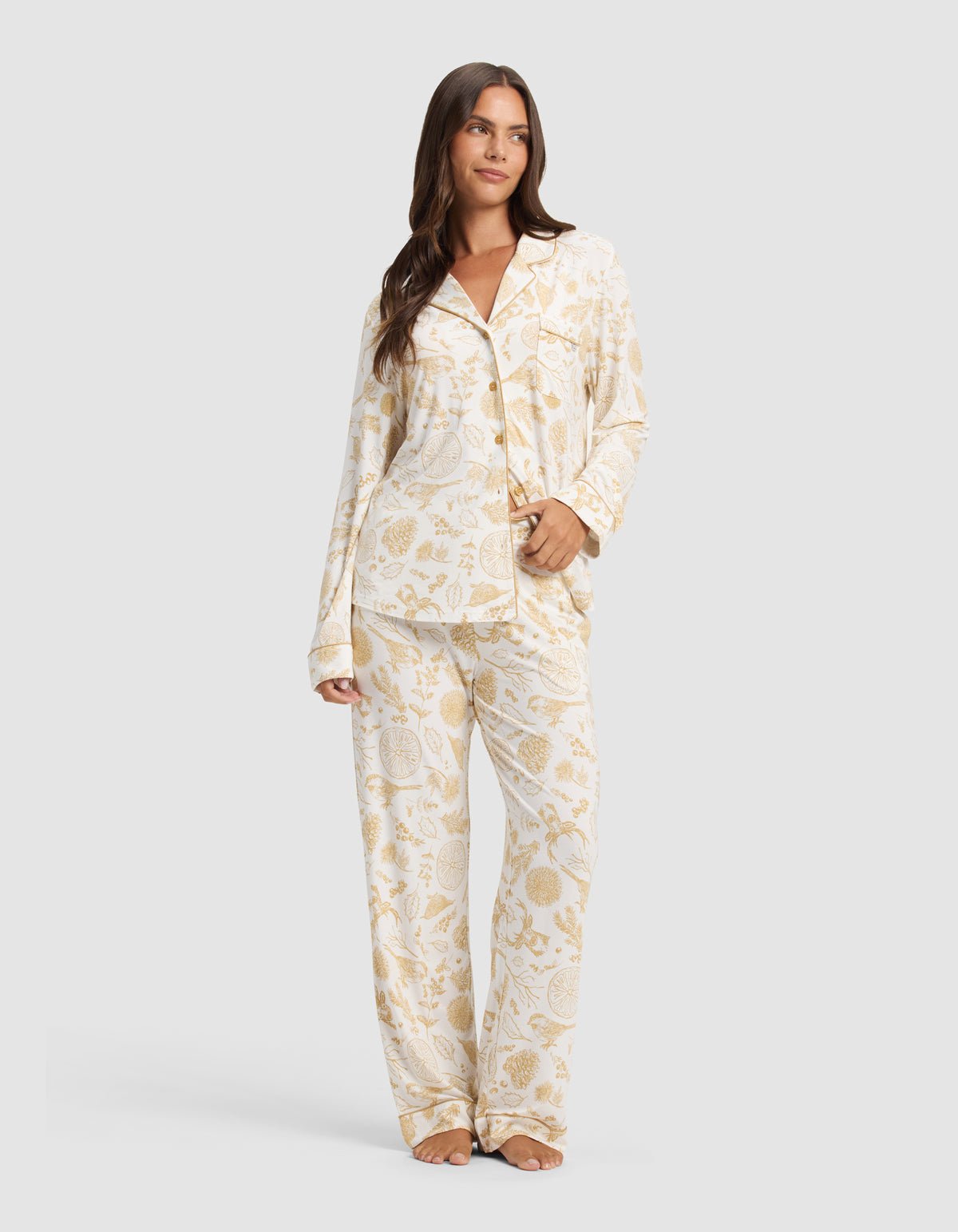 A woman stands barefoot against a light gray background wearing Cozy Earth's Women's Bamboo Stretch-Knit Pajama Pant in cream with a gold botanical print, paired with a matching long-sleeve button-up shirt.|Color:Winter Toile Dijon
