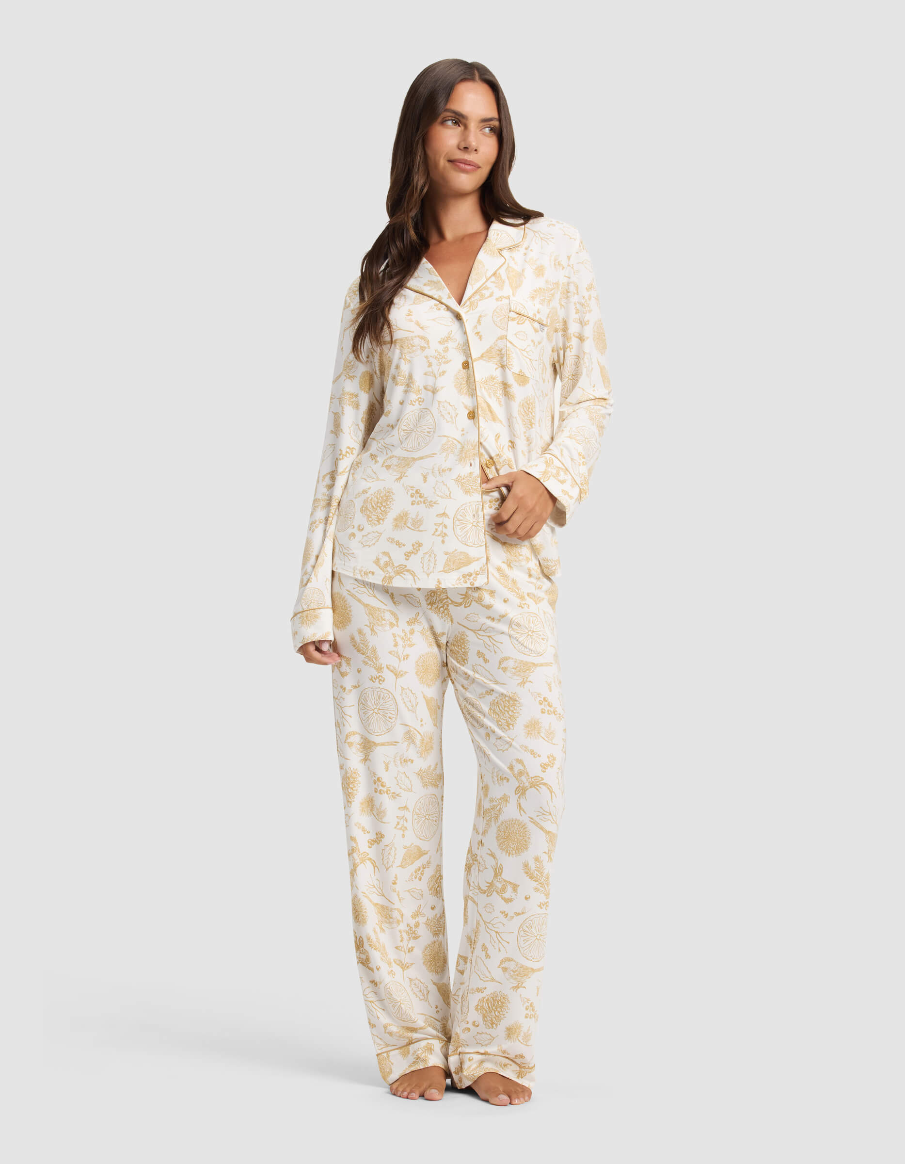 A woman with long brown hair stands barefoot against a light gray background, wearing Cozy Earth’s Women's Bamboo Stretch-Knit Pajama Pant in cream with a gold floral pattern, looking forward with a relaxed expression.