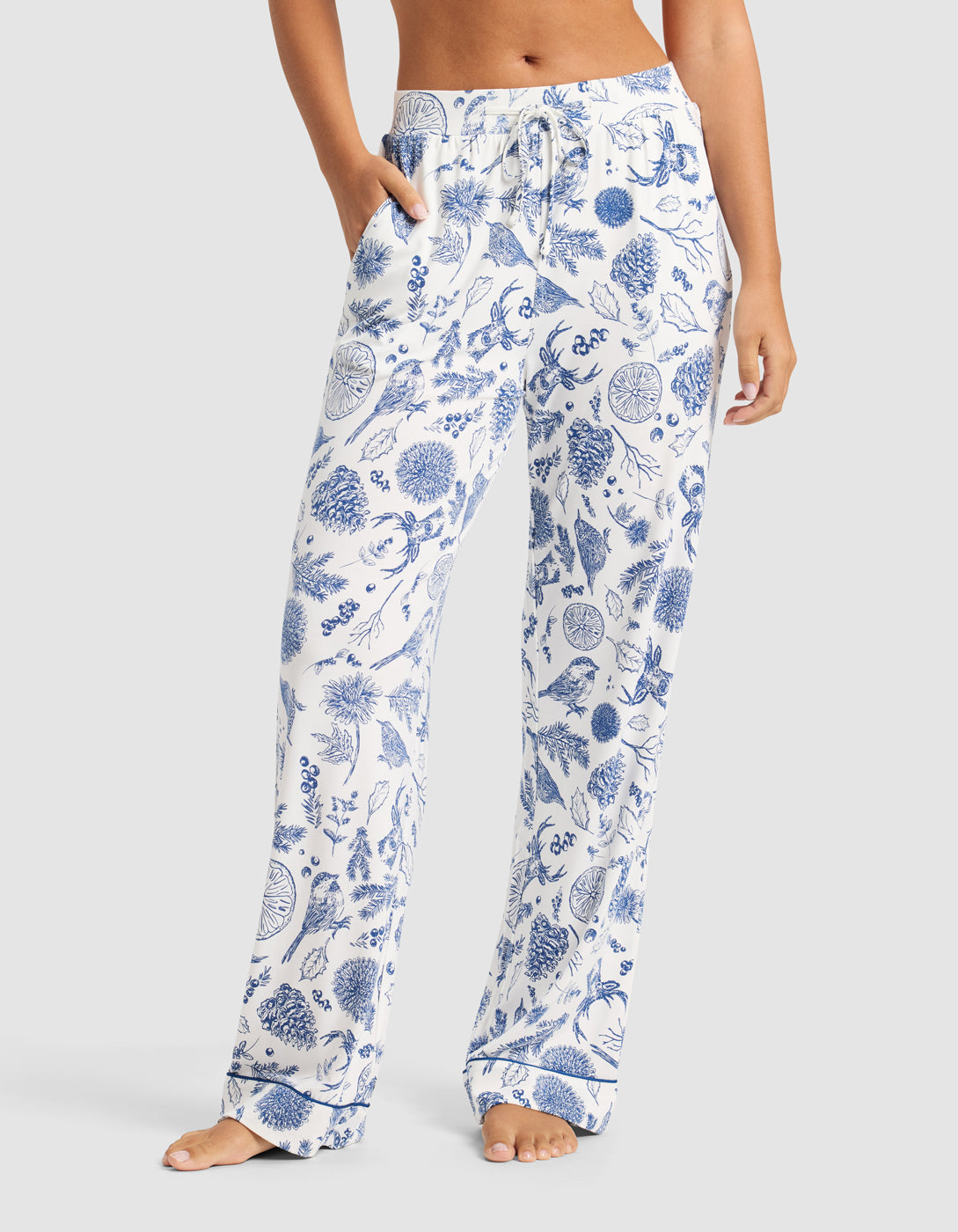 A woman wears Cozy Earth’s Women's Bamboo Stretch-Knit Pajama Pant in white with a blue botanical print, standing barefoot with hands in pockets against a plain background.