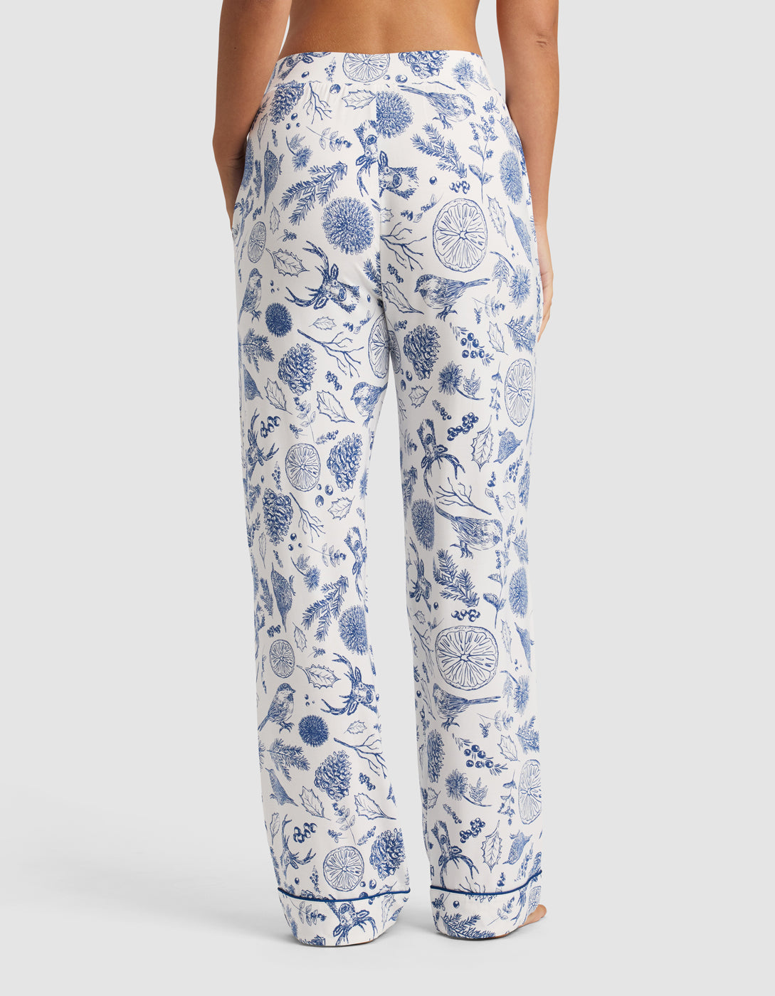A person stands with their back to the camera wearing Cozy Earth Women's Bamboo Stretch-Knit Pajama Pant in white with a blue floral print. The pants are loose-fitting with pockets, showing only the lower back and legs against a plain background.